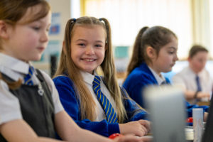 Three young girls are pictured sat at their desks, wearing their academy uniform. The girl in the centre is seen smiling brightly at the camera.