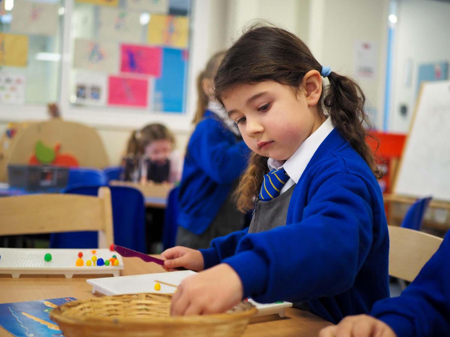 A young girl is seen wearing her academy uniform whilst sitting at a desk, interacting with different coloured objects.