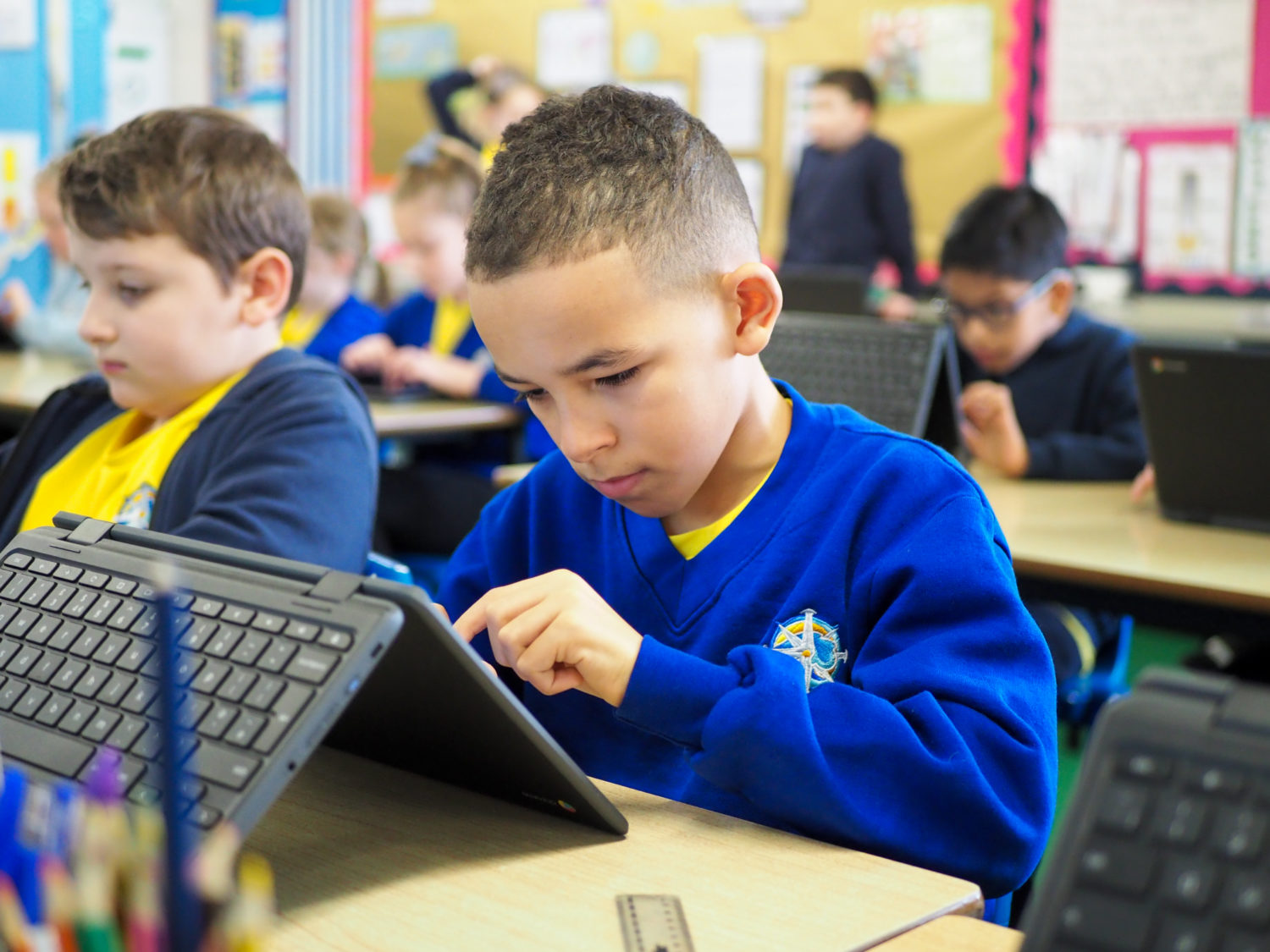 A young boy is seen sat at his desk using a tablet computer in a classroom.