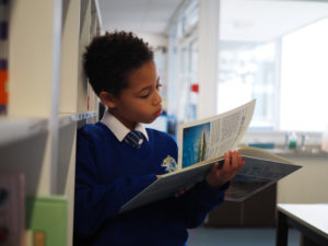 A young boy is shown wearing his academy uniform, standing reading a book in a corridor.