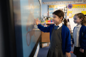 Two young girls are shown interacting with an Interactive Board. One of them is seen pointing at a section with her finger.