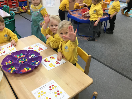 Nursery children sat at a table together waving at the camera.
