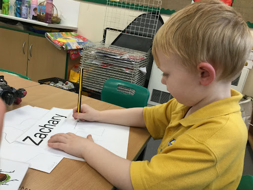 A Nursery child sat at a table writing on a sheet of paper.